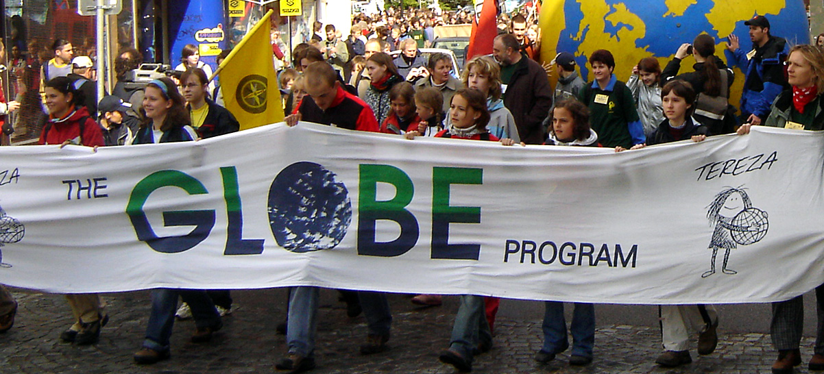 Several people in a large street participate in a parade. People hold yellow and blue flags. The people leading the parade hold a banner that reads, “The GLOBE Program."